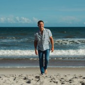 A man walking away from the sea on a beach wearing a blue printed shirt and blue jeans